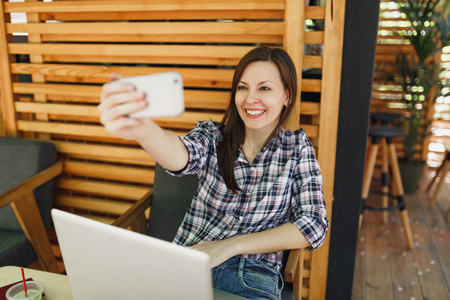 女士在木制户外街道夏季咖啡店，坐在笔记本电脑电脑上，在手机上自拍，在空闲时间放松。 移动办公室。 生活方式自由职业商业概念