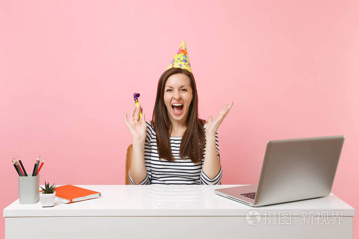喜出望外的女人戴着生日派对帽一边吹着烟斗尖叫庆祝一边坐在办公桌上