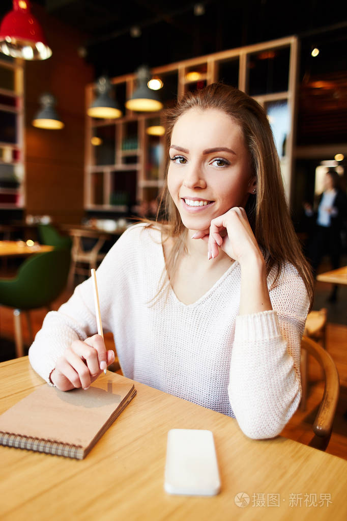 内容漂亮的女人坐在桌子旁,拿着电话和记事本,在咖啡馆里微笑着看镜头