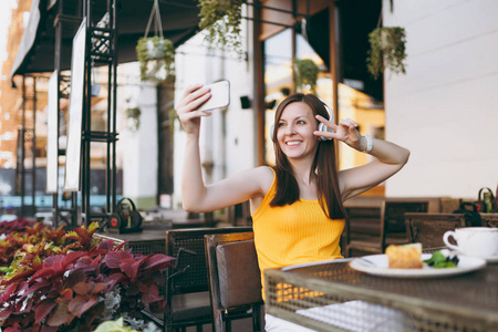 迷人的微笑女人在户外街头咖啡店咖啡馆坐在桌子旁听音乐耳机，在手机上自拍，在餐厅空闲时间放松。 生活方式休息概念
