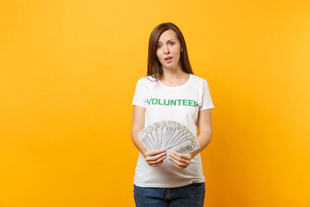 穿着白色T恤的女人，上面写着绿色标题，志愿者持有大量的美元，现金，孤立在黄色背景上。 自愿无偿援助慈善恩典工作概念
