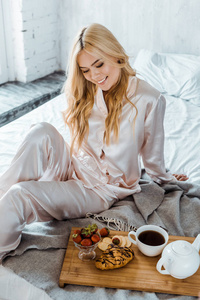 穿着睡衣微笑着迷人的女人看着床上木制托盘上的早餐