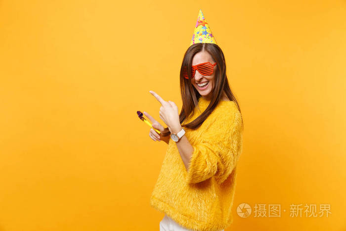 欢快的快乐女人戴着橙色滑稽的眼镜生日帽吹着烟斗食指指着旁边的复制