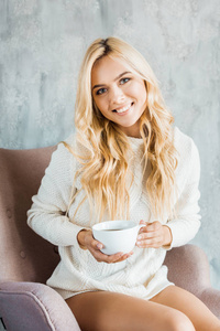 早上，穿着毛衣坐在扶手椅上在卧室里拿着一杯咖啡的迷人的微笑女人