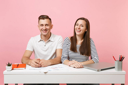 两个年轻的微笑商务女性男性同事坐在白色办公桌上工作，现代笔记本电脑被隔离在粉彩背景上。成就职业概念。复制空间广告青年合作