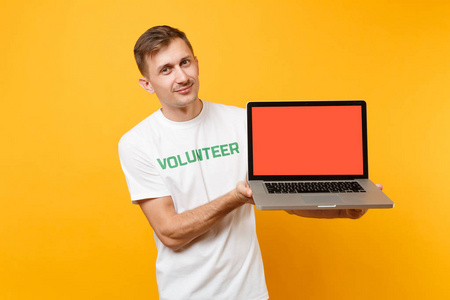 男子穿白色T恤，文字铭文，绿色标题，志愿者持有笔记本电脑，空白空屏幕隔离在黄色背景。 自愿无偿援助慈善恩典工作概念