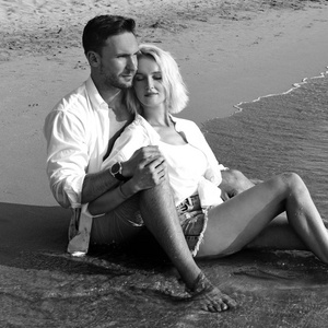 美丽的幸福夫妇相爱，躺在沙滩上，穿着白色的湿衬衫，爱着微笑。 英俊的留着胡须的男人和的金发美女穿着湿衣服在水中玩着有趣的欢笑
