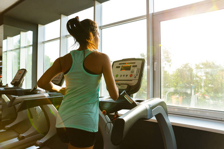年轻的运动女性在健身房窗户附近的跑步机上跑步