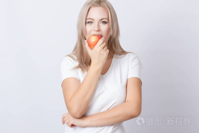 一位年轻漂亮的金发长发女子站在灰色背景上手里拿着苹果吃着苹果