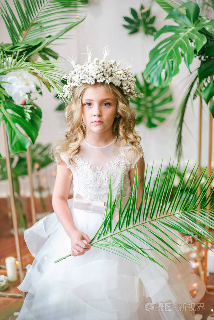 可爱的小女孩金色的卷发穿着白色的婚纱戴着热带树叶