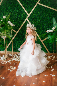 可爱的小女孩，金色的卷发，穿着白色的婚纱，戴着热带树叶白色玫瑰和兰花装饰的花圈