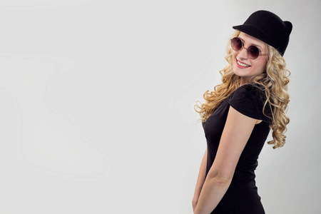 穿着黑色T恤和帽子，戴着太阳镜，摆着调情姿势的时髦金发女人的侧视图