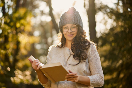 戴着帽子和眼镜的女人手里拿着一本书在秋天的森林里。