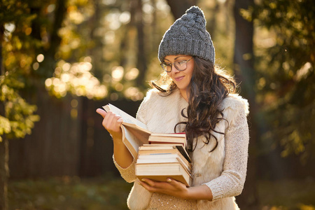 戴着帽子的女人拿着一堆书在秋季森林的背景上。