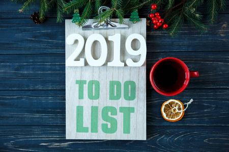 在木制垫白色号码2019做清单与红色咖啡或茶和冷杉树装饰在灰色木桌上。 新年目标列出了圣诞节概念上要做的事情。 平躺着有共空间。