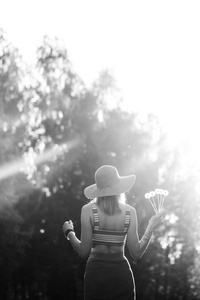 黑白图像。 女孩在公园户外散步的剪影。 阳光明媚的夏季生活方式概念。 穿裙子和帽子的女人在田野里用蒲公英。 光效应和文本的共空间