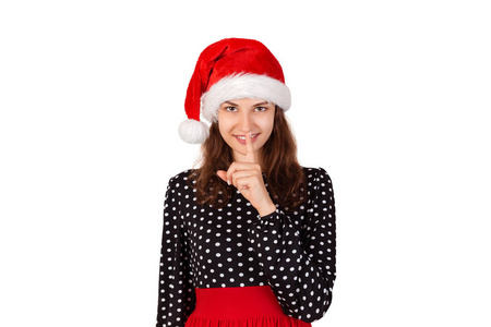 女人睁大眼睛，用手指在嘴唇上要求沉默或保密。 情感女孩在圣诞老人圣诞帽隔离在白色背景。 假日概念。
