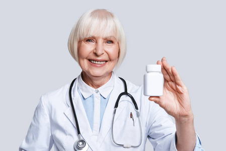 穿着实验室外套拿着药瓶子站在灰色背景下微笑的漂亮老妇人