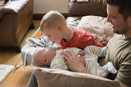 父亲坐在沙发上，他的小男孩站在他的腿上，他的儿子试图用奶嘴来安慰他的弟弟。
