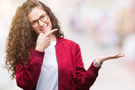 美丽的黑发卷发年轻女孩穿着夹克和眼镜在孤立的背景上惊讶和微笑的相机，同时呈现手和手指。