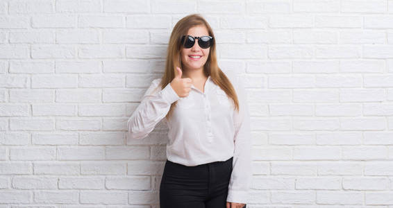 年轻的成年商务女性站在白色的砖墙上戴着太阳镜，开心地大笑着做OK标志，用手指竖起大拇指，非常棒的标志