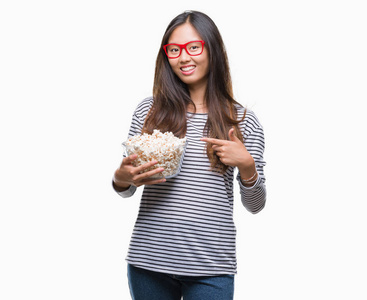 年轻的亚裔妇女在孤立的背景下吃爆米花，非常高兴地用手和手指指着。