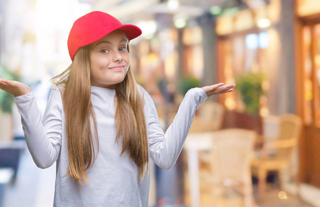 年轻漂亮的女孩戴着红色的帽子，背景模糊，表情混乱，举起手臂和手。 怀疑概念。