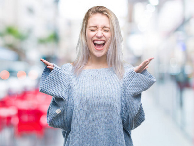 年轻的金发女人穿着冬天的毛衣，在孤立的背景上庆祝疯狂和疯狂的成功，手臂抬起和闭上的眼睛尖叫兴奋。 赢家概念