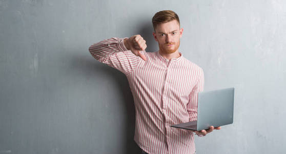 年轻的红头发的男人，穿着灰色的灰色衣服，拿着电脑笔记本电脑，脸上带着愤怒的，表示不喜欢，拇指向下拒绝的概念