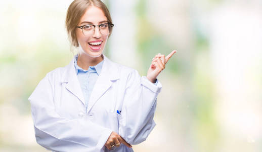 美丽的年轻金发医生女人穿着白色外套，在孤立的背景上，脸上挂着一个大大的微笑，用手和手指指向一边，看着相机。