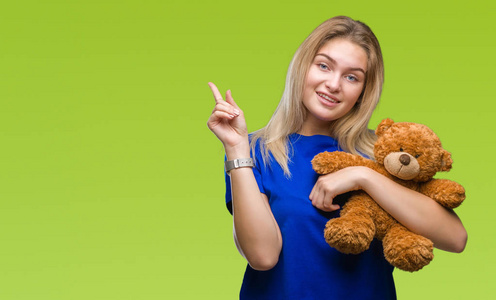 年轻的白种人女人抱着可爱的泰迪熊在孤立的背景上，非常高兴地用手和手指指向一边