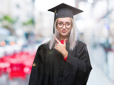 年轻的金发女人穿着研究生制服，在孤立的背景上欢快地笑着，脸上带着微笑，用手和手指指向一边，脸上带着快乐和自然的表情看着相机。