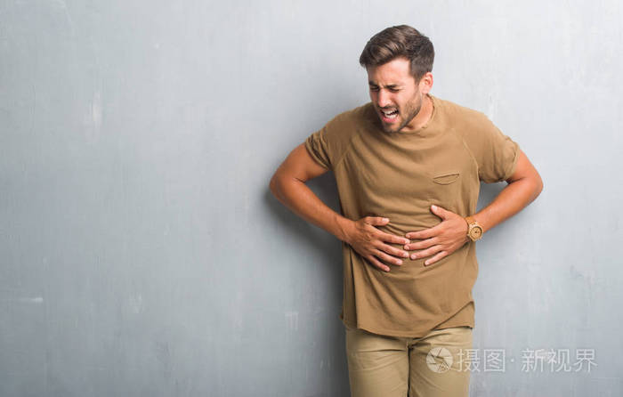英俊的年轻人在灰色的墙壁上,手在肚子上,因为消化不良,痛苦的疾病