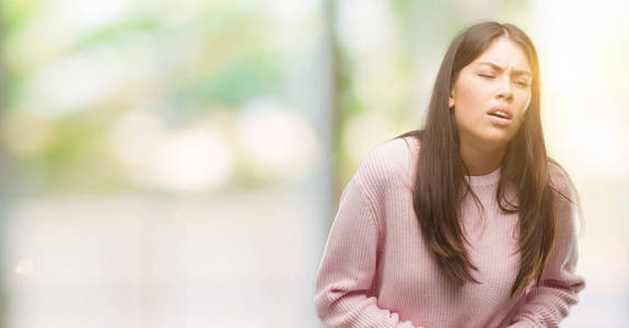 年轻漂亮的西班牙裔女人穿着毛衣，手放在肚子上，因为消化不良，痛苦的疾病，感觉不舒服。 疼痛的概念。