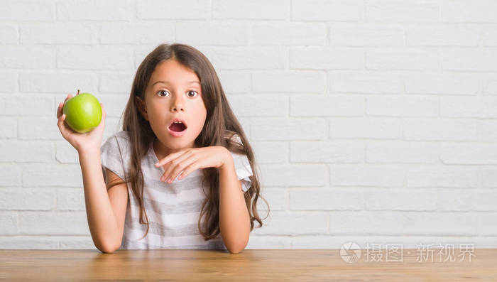 年轻的西班牙裔小孩坐在桌子上吃着新鲜的绿苹果,吓得满脸惊讶,恐惧