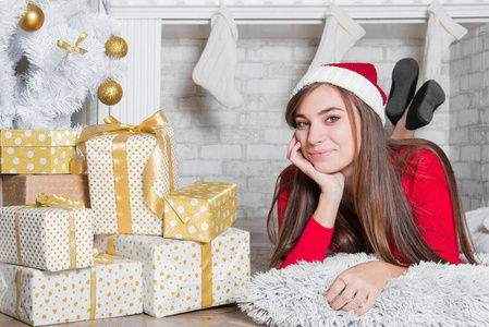 年轻迷人的黑发女人靠近圣诞节壁炉在舒适的家庭内部。 戴着红色圣诞帽的十几岁女孩。 寒假概念。 白色漂亮的装饰。 圣诞客厅里有趣的