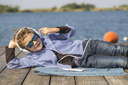 一个带太阳镜和耳机的小可爱男孩的肖像。 他躺在河边听音乐，享受着美丽的晴天。 他是一个非常现代和有魅力的男孩