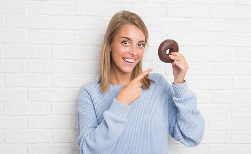 美丽的年轻女子在白色砖墙上吃巧克力甜甜圈，用手和手指指着非常高兴