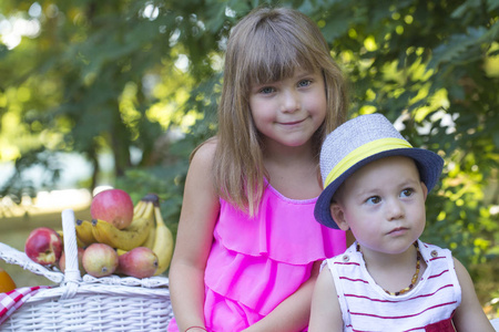小可爱的男孩和女孩坐在草地上的毯子上玩。 女孩拥抱了男孩。 在男孩和女孩附近有一个野餐篮，里面有果汁和很多水果。
