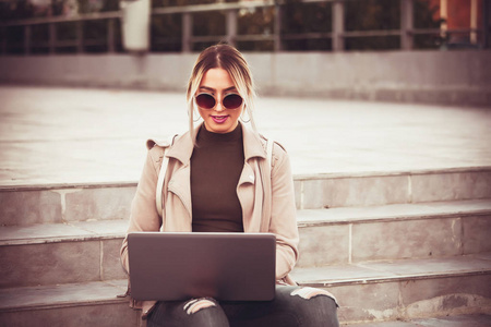 幸福的年轻女人坐在外面的楼梯上，在笔记本电脑上工作。 无线互联网远程工作自由职业者打印一个信息通信在社交网络。