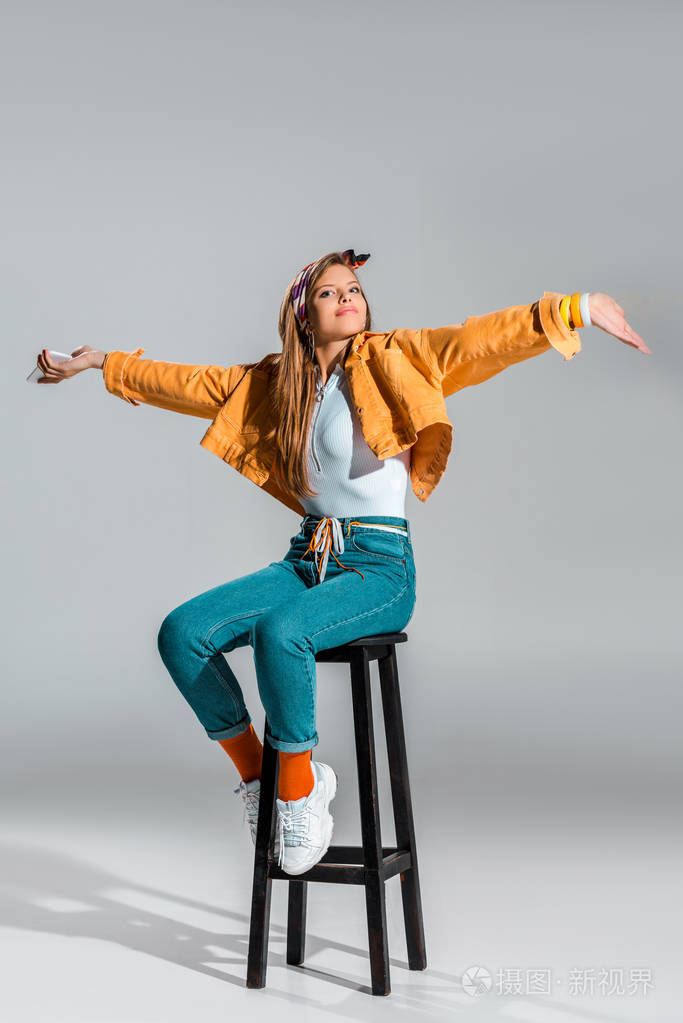 美丽的女孩伸出双手拿着智能手机坐在灰色的凳子上