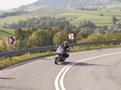 大胡子摩托车手戴着头盔太阳镜和黑色皮衣，驾驶自行车沿着空旷的道路在明亮的夏日，雾蒙蒙的和遥远的青山背景。