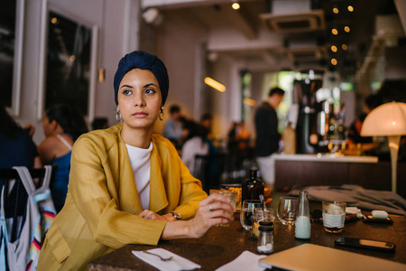年轻优雅的穆斯林马来女人在咖啡馆里喝咖啡。 她穿着时尚的衣服，头巾头巾和耳环。