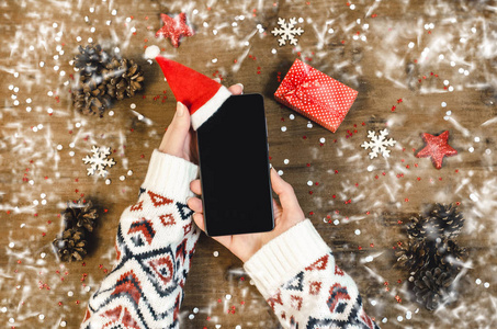 女性手拿着智能手机，在木制背景上展示OLED，圣诞礼物雪花和雪。 新年快乐帽子圣诞老人在手机平躺构图顶部视图。