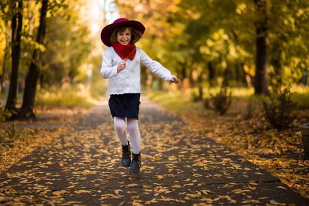 可爱的小女孩戴着红帽子走在秋天的美丽公园里