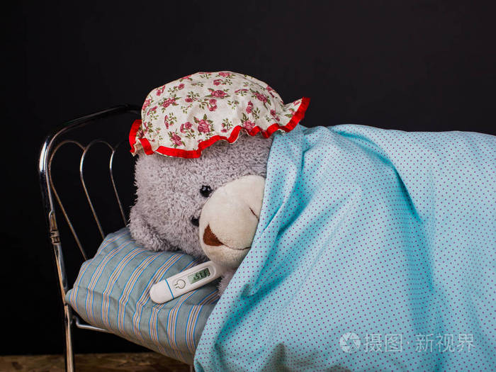 一只生病的孤独的泰迪熊躺在床上带着体温计躺在一个黑色背景的卧室里