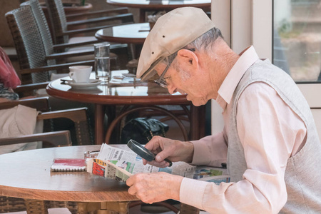 戴眼镜的老人在咖啡馆的桌子旁用放大镜看报纸照片