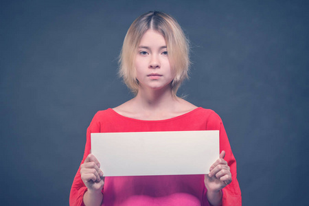 金发女孩少年穿着一件红色衬衫，在灰色背景上举着一张空白的白色海报。 色调