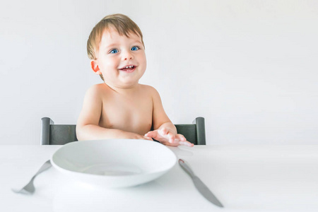 笑着，蓝眼睛的金发小男孩坐在桌子旁，手里拿着一把白色的盘子叉子和刀子