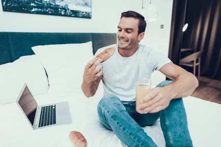 快乐的人在床上工作和吃早餐。 白种人的肖像微笑者穿着浅色T恤和蓝色牛仔裤，使用灰色笔记本电脑，在现代公寓里吃三明治和喝新鲜果汁。